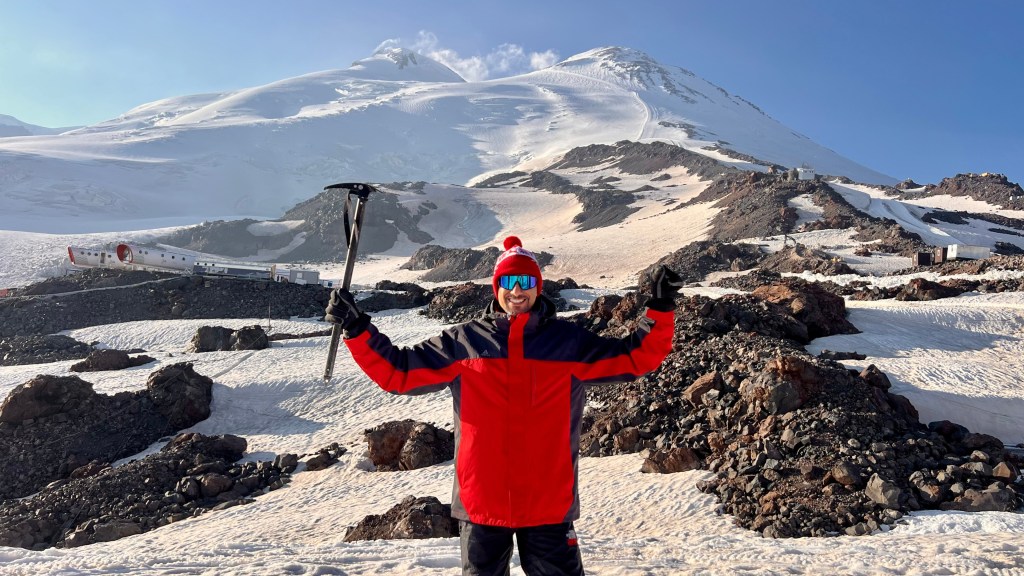Mohamed Isa raising an ice axe on the snow below the twin peaks of Mount Elbrus